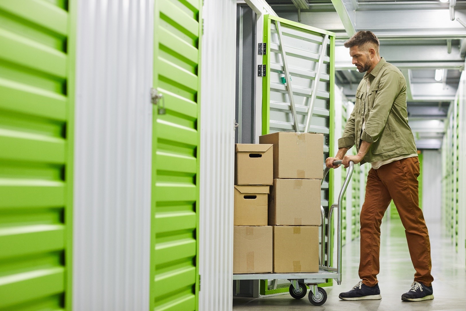 A man wheeling some boxes into a storage unit. The facility is grey and clean, with lime green doors.
