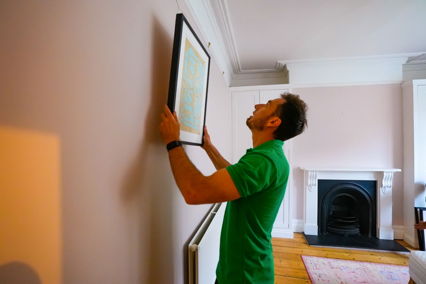 A Man in a green shirt hangs up a photo in an empty, new house. A rug is on the wooden floor, and a fireplace is in the background.